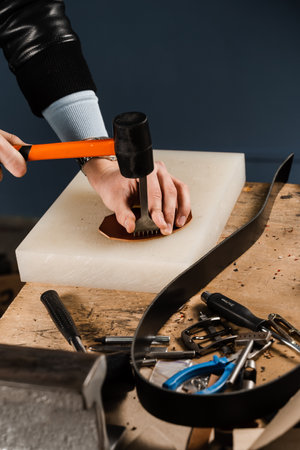Craftsman is punching genuine leather using line punch and hammer for creation natural leather products. Tools of craftsman for belts creation. Equipment for genuine leather production in workshopの写真素材