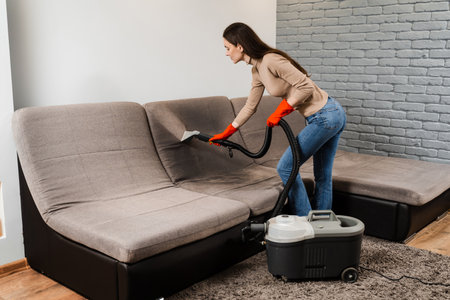 Cleaner girl is cleaning couch with extraction machine for dry clean upholstered furniture. Housekeeper is extracting dirt from upholstered sofa using dry cleaning extractor machineの写真素材