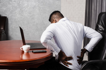 African man feels back pain after working for long hours with laptop online in office. Cervical spine osteochondrosis is radicular syndromes of african american manの写真素材