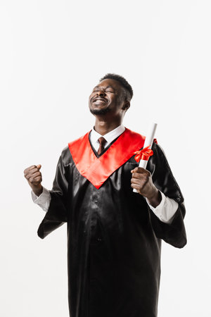 Graduate african man is graduating college and celebrating academic achievement. Happy african student in black graduation gown and cap raises masters degree diploma above head on white backgroundの写真素材