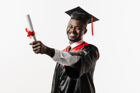 Happy african student in black graduation gown and cap raises masters degree diploma above head on white background. Graduate african man is graduating college and celebrating academic achievementの写真素材