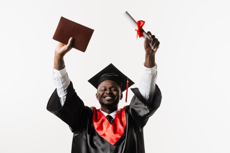 Happy african student in black graduation gown and cap raises masters degree diploma above head on white background. Graduate african man is graduating college and celebrating academic achievementの写真素材