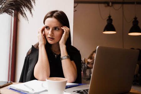 Sad girl with laptop got migraine touches her head because of pain. Woman manager with strong headache working on laptop online remotely in loft cafeの写真素材