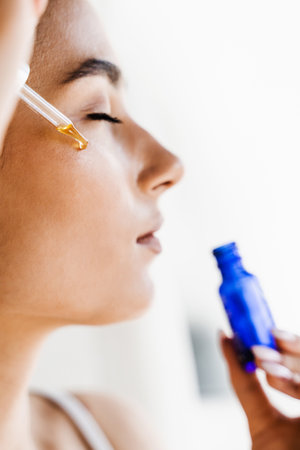 Young woman is applying moisturizing serum on her face. Attractive girl drops essential oil on...