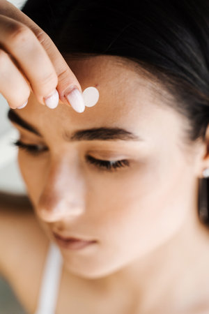 Young woman using acne patches for treatment of pimple and rosacea close-up. Facial rejuvenation cleansing cosmetology. Girl with acne stick round acne patch on her cheekの写真素材