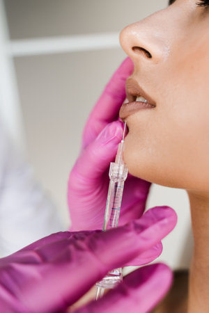 Close-up lips augmentation injections in lips for attractive girl. Cosmetologist injecting...