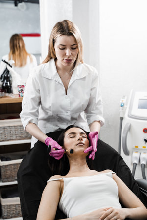 Cosmetologist using stone roller for face massage for girl patient. Facial massage with cosmetics roller in cosmetology clinic. Process of face massage with black stone roller close-upの写真素材