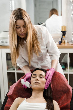 Cosmetologist is doing ultrasonic peeling skin with skin scrubber for girl patient at cosmetology clinic. Ultrasound facial peeling using scrubberの写真素材