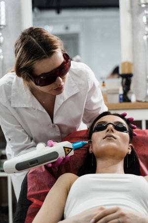 Cosmetologist cleans face of girl from pigmentation using laser. Hardware treatment. Laser removal of blood vessels on face of girl in dermatological clinicの写真素材