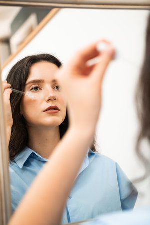 Girl with hyaluronic acid or serum pipette in hands is looking in the mirror. Young woman is...