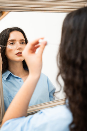 Attractive girl drops essential oil on facial skin in the mirror at home. Young woman is applying moisturizing serum on her faceの写真素材