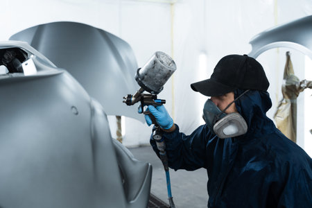 Mechanic from vehicle maintenance service is spreading paint on car using dark spray and paint gun....