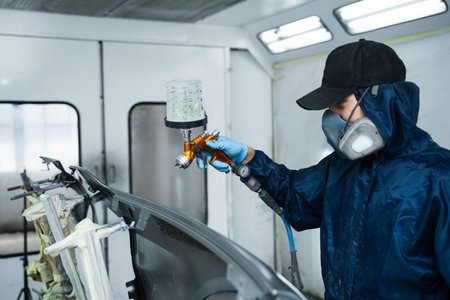 Car painter is finishing his paintwork on vehicle by applying lacquer with paint sprayer. Using LVLP spray gun to spread lacquer on metalの写真素材