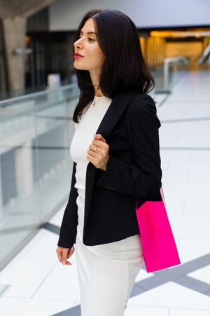 Enjoying shopping and buying new clothes as hobby. Happy attractive woman is going shopping on Black Friday. Woman in white dress and black jacket is walking in city storeの写真素材