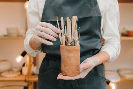 Clay crafting tools for ceramists. Potter in black apron holds a bunch of instruments for sculpting pottery in ceramic mugの写真素材