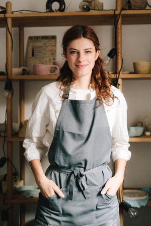 Happy young potter stands and smiling with shelves filled with earthenware in the background in her...