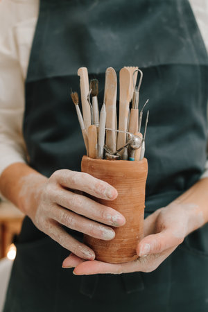 Potter in black apron holds a bunch of instruments for sculpting pottery in ceramic container. Clay crafting tools for ceramistsの写真素材