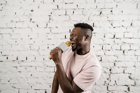 Handsome and cheerful singer is performing his song. Recording the audio in the studio. Young african man is enjoying singing in the microphone.の写真素材