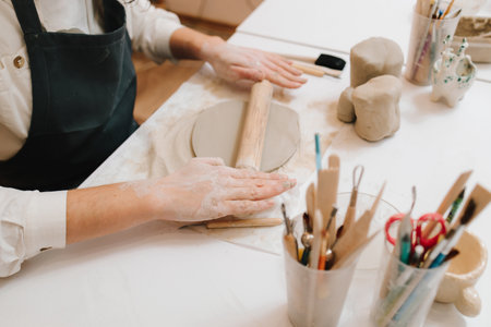 Woman artisan carefully rolls clay with pottery tools in the ceramic studio. Worker molds clay with...