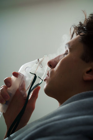 Sick man wrapped in a blanket is doing inhalation by nebulizer for the cough treatment. Patient is...