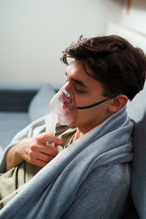 Sick man wrapped in a blanket is doing inhalation by nebulizer for the cough treatment. Patient is using inhaler for home treatmentの写真素材