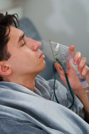 Inhaling air by nebulizer mask for cough therapy at home. Sick man has respiratory treatment by...