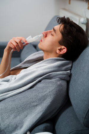 Young man with asthma attack breathing with inhaler in the bed. Doing nose inhalation with nebulizer at homeの写真素材