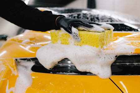 Hand washing with yellow sponge and foam for sports car body in garage. Car washer doing manual foam washing in auto detailing serviceの写真素材