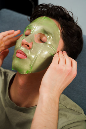 Relaxed man with hydrating face mask at home. Man is applying facial mask for rejuvenate complexion, skincare conceptの写真素材