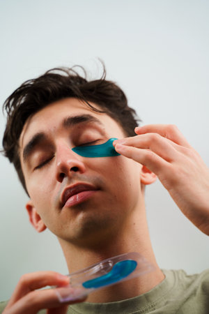 Male skincare routine at home. Young man with sensitive skin wears hydrogel patches under his eyesの写真素材