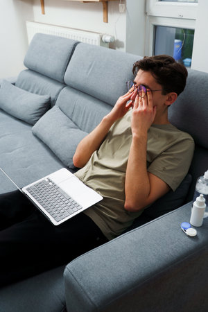 Tired IT specialist with headache in front of laptop. Eye tiredness from working remotely at homeの写真素材