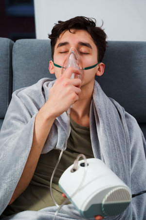 Sick man wrapped in a blanket is doing inhalation by nebulizer for the cough treatment. Patient is using inhaler for home treatmentの写真素材