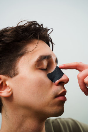 Man with charcoal nose strip for pores cleaning. Young guy applying charcoal nose strip for deep nose pores cleaning. Male beauty and skincare conceptの写真素材