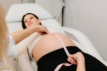 Gynecologist checks size of the woman's belly in the clinic. Gynecologist measures pregnant belly with measuring tape to follow the growth of the baby on the appointmentの写真素材