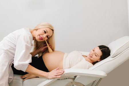 Gynecologist leans a fetal stethoscope to a pregnant woman's belly and listens to the baby's heartbeat. Gynecologist makes examination of pregnant woman's belly on the appointment at hospitalの写真素材