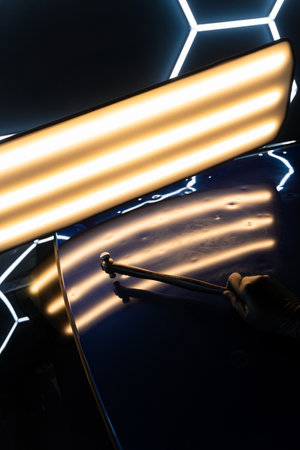 PDR technician holds a specialized hammer against a reflection light board to check dent correction on the hood. Accurate paintless car dent removalの写真素材