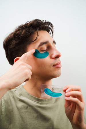 Male skincare routine at home. Young man with sensitive skin wears hydrogel patches under his eyesの写真素材