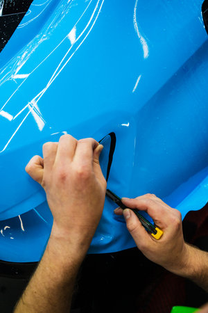 Cutting blue paint protection film with blade during detailing job ensures precision edges. Detailing technician trims blue PPF on a car panel for perfect contour fit. Cutting blue PPF with bladeの写真素材