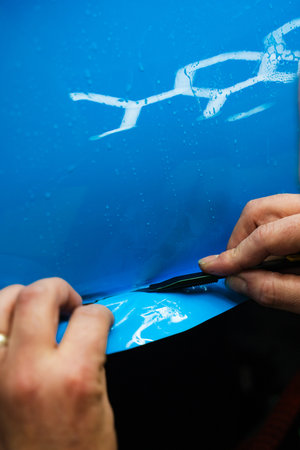 Detailing technician trims blue PPF on a car panel for perfect contour fit. Cutting blue PPF with blade. Cutting blue paint protection film with blade during detailing job ensures precision edges.の写真素材