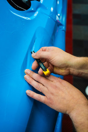 Detailing technician trims blue PPF on a car panel for perfect contour fit. Cutting blue PPF with blade. Cutting blue paint protection film with blade during detailing job ensures precision edgesの写真素材