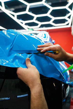 Cutting blue PPF with blade. Cutting blue paint protection film with blade during detailing job ensures precision edges. Detailing technician trims blue PPF on a car panel for perfect contour fitの写真素材