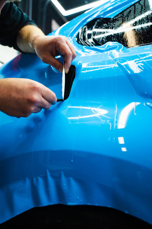 Detailing technician trims blue PPF on a car panel for perfect contour fit. Cutting blue PPF with blade. Cutting blue paint protection film with blade during detailing job ensures precision edgesの写真素材