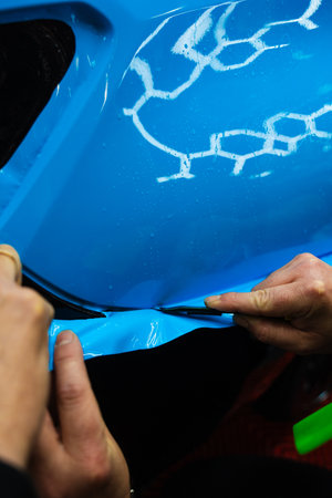 Cutting blue PPF with blade. Cutting blue paint protection film with blade during detailing job ensures precision edges. Detailing technician trims blue PPF on a car panel for perfect contour fitの写真素材