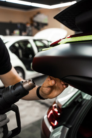 Car Vinyl Wrapping with Heat Gun. Applying black vinyl wrap to car with heat gun ensures precise adhesion and flawless surface coverage. Heat gun used in vinyl wrapping activates adhesiveの写真素材