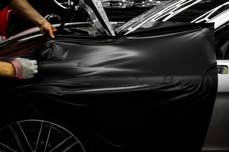 Car wrapping process using black matte vinyl stretched into place. Careful application and tensioning of black vinyl avoids surface damage or bubbling. Matte black vinyl is applied across car hoodの写真素材
