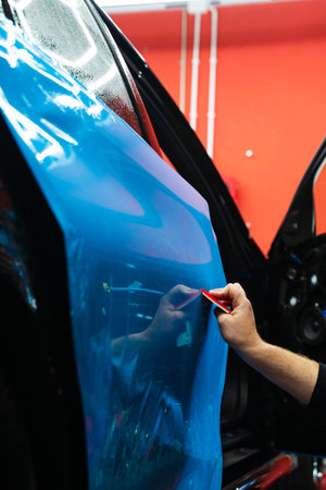 Detailing expert uses red squeegee wrapper to flatten blue PPF wrap on car surface. Vehicle wrap detailing involves squeegee strokes for seamless, wrinkle-free adhesionの写真素材