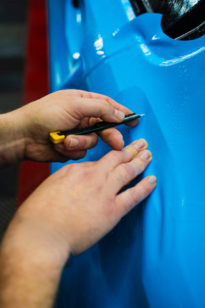 Detailing technician trims blue PPF on a car panel for perfect contour fit. Cutting blue PPF with blade. Cutting blue paint protection film with blade during detailing job ensures precision edgesの写真素材