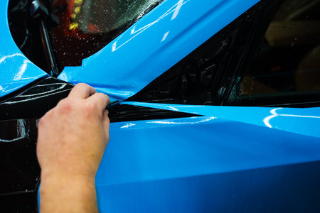 Detailing technician trims blue PPF on a car panel for perfect contour fit. Cutting blue PPF with blade. Cutting blue paint protection film with blade during detailing job ensures precision edges.の写真素材