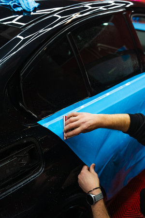 Detailing expert uses red squeegee wrapper to flatten blue PPF wrap on car surface. Vehicle wrap detailing involves squeegee strokes for seamless, wrinkle-free adhesionの写真素材
