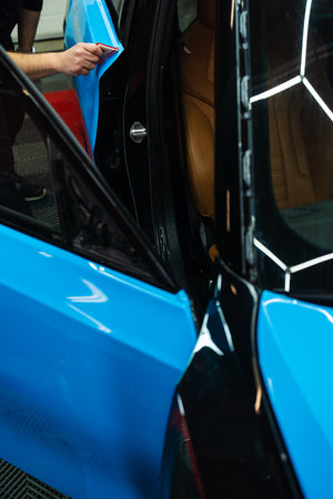 Detailing expert uses red squeegee wrapper to flatten blue PPF wrap on car surface. Vehicle wrap detailing involves squeegee strokes for seamless, wrinkle-free adhesionの写真素材